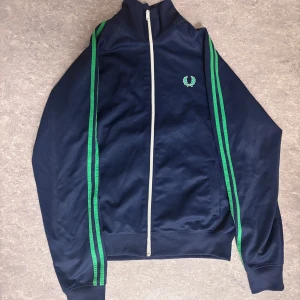  Tracktop från Fred Perry - Säljer en snygg blå tracktop från Fred Perry med gröna detaljer och dragkedja. Skick 9/10