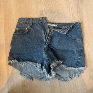 Blå jeansshorts från NA-KD - Snygga blå jeansshorts från NA-KD med fransiga kanter och klassisk femficksdesign. Perfekta för en avslappnad sommarlook. De har en knapp och dragkedja framtill.