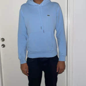 Ljusblå hoodie från Lacoste - Säljer en stilren ljusblå hoodie från Lacoste med klassisk krokodil-logga på bröstet. Tröjan har en känguruficka och justerbar huva med dragsko. Perfekt för en avslappnad look.
