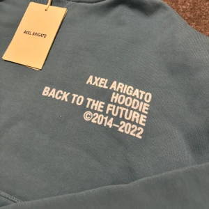 Grön/blå hoodie från Axel Arigato - Såå snygg grön hoodie från Axel Arigato med texten 'Back to the Future' tryckt på bröstet. Den har en klassisk känguruficka och justerbar huva med dragsko. Perfekt för en avslappnad stil. Aldrig använd på grund av att jag inte fått något tillfälle till det. XS men passar S också då jag skulle säga att den är stor i storleken.