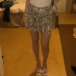 Mönstrad kjol från Gina Tricot - Snygg kjol från Gina Tricot i beige och vitt med blommigt mönster. Kjolen har en knytning framtill som ger en fin detalj och en omlott-look. Somrig och fin!!