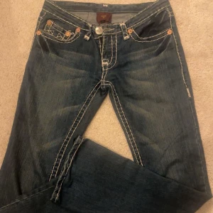 Blå jeans från True Religion - Snygga blå jeans från True Religion med distinkta vita sömmar och klassisk femficksdesign. Byxorna har en lågmidjad passform och är dekorerade med märkets ikoniska broderi på bakfickorna. ‼️kan gå ner i pris vid snabb affär‼️