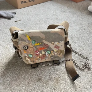 Zadig & Voltaire väska - Säljer en cool axelväska i camouflage med unika patches och kedjedetaljer. Väskan har texten 'Revolution' på axelremmen och flera färgglada motiv som stjärnor och blixtar. Perfekt för att ge din outfit en edgy touch.