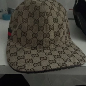 Beige keps med monogrammönster - Snygg beige keps från Gucci med det ikoniska monogrammönstret. Kepsen har en justerbar rem baktill och detaljer i rött och grönt på sidan. Perfekt för att ge din outfit en lyxig touch.