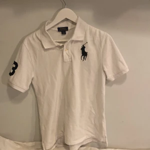 Vit pikétröja från Ralph Lauren 14-16 år - Säljer en klassisk vit pikétröja från Ralph Lauren med korta ärmar. Tröjan har den ikoniska broderade loggan på bröstet och en stor siffra 3 på ärmen. Perfekt för en stilren och sportig look.