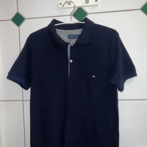 Mörkblå pikétröja från Tommy Hilfiger - Snygg mörkblå pikétröja från Tommy Hilfiger med klassisk krage och knappdetalj. Den har ett diskret märke på bröstet och ett stilrent rutigt mönster på insidan av kragen. Perfekt för en avslappnad men stilren look.