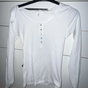 Vit långärmad topp med knappar - Säljer en stilren vit långärmad topp med knappar framtill. Perfekt för en enkel och klassisk look. Toppen har en rund halsringning och är tillverkad i ett mjukt material.