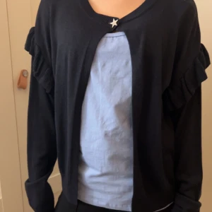 Mörk blå kofta med volangdetaljer - Snygg mörk blå kofta med volangdetaljer på ärmarna och en stjärnformad knapp framtill. Perfekt för att lägga till en touch av stil till din outfit.