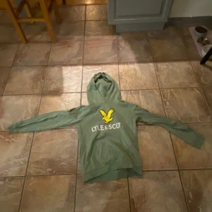 Grön hoodie från Lyle & Scott - Säljer en grön hoodie från Lyle & Scott med deras ikoniska gula örnlogga på bröstet. Tröjan har långa ärmar och en bekväm huva, perfekt för en avslappnad stil.