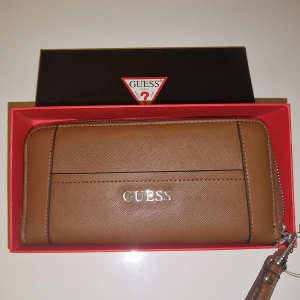 Plånbok från Guess - Snygg plånbok från Guess med dragkedja och avtagbar handledsrem. Tillverkad i syntetmaterial med stilren design och Guess-logga i silver på framsidan. Matchande väska säljes. Garderobsrensning så kika gärna in övriga auktioner. 