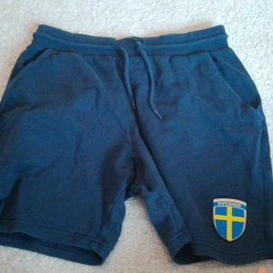 Blå supporter shorts med Sverige-emblem - Snygga blå supporter shorts med Sverige-emblem på benet. Perfekta för att visa ditt stöd under matcherna. De har en bekväm passform med dragsko i midjan och praktiska fickor.