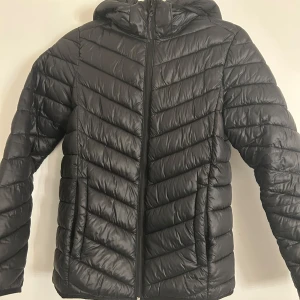 Svart pufferjacka med huva - Snygg svart pufferjacka med huva och dragkedja. Jackan har ett quiltat mönster och är perfekt för kyligare dagar. Den har långa ärmar och en stilren design.