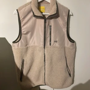 Beige fleeceväst från Rotation - Säljer en beige fleeceväst från Rotation i storlek medium. Västen har en dragkedja framtill och två fickor med dragkedjor. Perfekt för kyliga dagar.