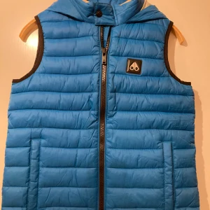 Blå väst  från Moose Knuckles - Snygg, blå,  tunn väst för barn från Moose Knuckles med dragkedja och huva (ej avtagbar). Perfekt för mindre kyliga dagar. Västen har ett stilrent märkeslogga  på  bröstet. Fickorna på sidorna är utan dragkedja. Rymliga fickor på insidan. Liten i storleken, L motsvarar 157-165 cm men skulle säga att den motsvarar snarare  150-160cm.  Använd  endast 3-5 gånger som i princip i nyskick. 