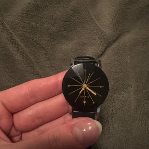 Svart klocka med guld detaljer - Stilren svart klocka med guldfärgade visare och detaljer. Klockan har en svart urtavla och ett svart armband i skinn. Perfekt för en elegant look.