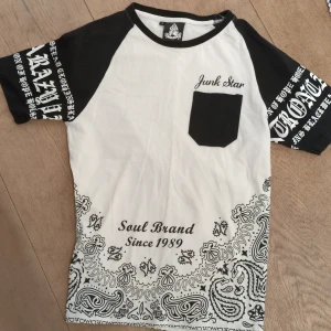 Vit och svart t-shirt från Junk Star - Snygg vit och svart t-shirt från Junk Star med tryck 'Soul Brand Since 1989'. Den har korta ärmar med textdetaljer och ett svart bröstficka. Bandana-mönster längst ner ger en unik touch.