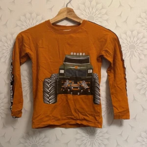 Orange långärmad tröja med monstertruckmotiv - Cool orange tröja med ett stort monstertrucktryck på framsidan. Långärmad design med textdetaljer längs ärmarna. Perfekt för den som älskar bilar och äventyr!