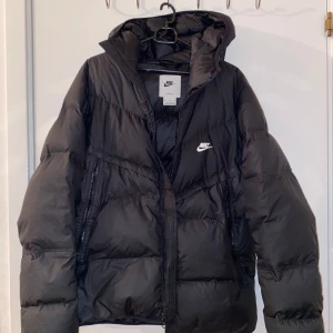 Svart dunjacka från Nike - Säljer en svart dunjacka från Nike med Storm-FIT teknologi. Jackan har en dragkedja framtill och en huva för extra skydd mot väder och vind. Perfekt för kyliga dagar. Köpte den för 2200kr men säljer den för 1500kr, pris kan diskuteras i chatten 