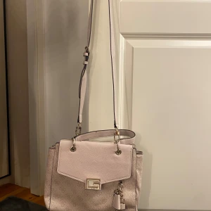 Ljusrosa axelväska från Guess - Snygg ljusrosa axelväska från Guess med justerbar axelrem och dekorativ tofs. Väskan har ett subtilt mönster och en praktisk dragkedja på baksidan. Perfekt för att bära dina dagliga nödvändigheter med stil. Helt oanvänd och legat länge i garderoben.