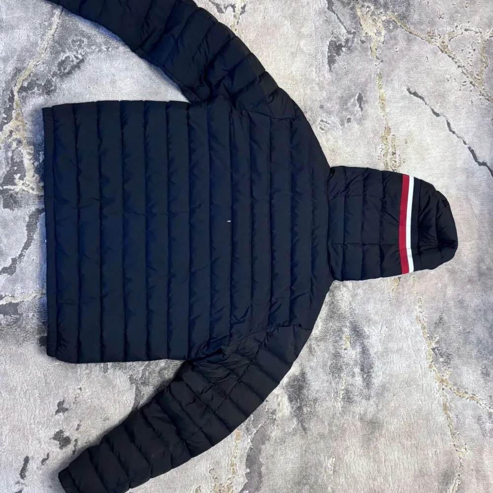 Snygg svart dunjacka från Moncler med huva och dragkedja. Jackan har ett stilrent mönster med ränder i rött och vitt på huvan. Perfekt för kyliga dagar.. Takit.