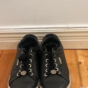 Svarta sneakers från Guess - Snygga svarta sneakers från Guess med snörning och metalliska detaljer. Perfekta för en stilren look. Märkeslogga på sidan och hälen ger en exklusiv touch. Skorna är lite slitna och smutsiga men det går att rensa. 