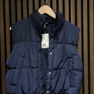 Mörkblå dunväst med knappar - Snygg mörkblå dunväst med knappar framtill. Perfekt för kyligare dagar och enkel att matcha med olika outfits. Västen har en stilren design och praktiska fickor.