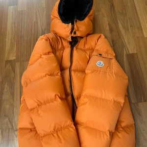 Orange dunjacka från Moncler - Snygg orange dunjacka från Moncler med huva och dragkedja. Jackan har två framfickor med dragkedjor och en logotyp på ärmen. Perfekt för kalla dagar med sin pufferdesign.