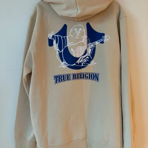 Beige hoodie från True Religion - Säljer en beige hoodie från True Religion med dragkedja och en stor logga på ryggen. Den har en justerbar huva och fickor framtill. Perfekt för en avslappnad stil. Helt ny med tag. Köpt i USA i december. 