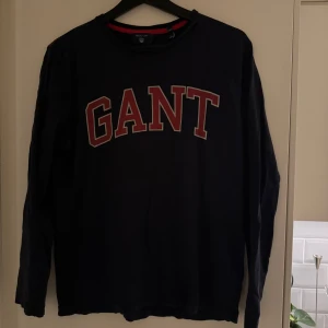 Marinblå långärmad tröja från GANT - Snygg marinblå långärmad tröja från GANT med röd och vit logga på bröstet. 