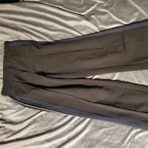 Svarta byxor från Monki - Snygga svarta byxor från Monki med elastisk midja och raka ben. Perfekta för en stilren look. Byxorna har en diskret vit rand längs sidorna som ger en sportig touch.