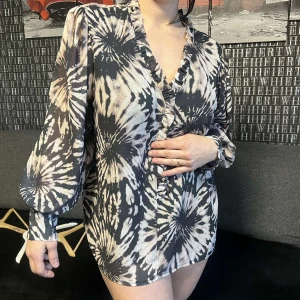 Svart och beige mönstrad klänning - Trendig klänning med svart och beige batikmönster. Den har en v-ringad design och långa puffärmar som ger en bohemisk känsla. Perfekt för en avslappnad stil.