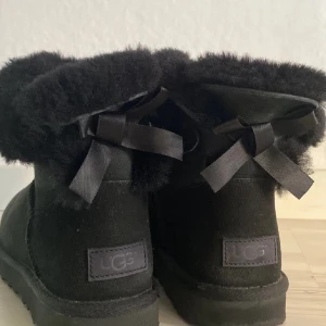 Svarta vinterskor UGGS - Säljer ett par svarta vinterskor UGGS med mjukt foder och dekorativa rosetter baktill. Perfekta för kalla dagar då dom är väldigt varma. De har en defekt som ett  hål (se sista bilden ) därav priset