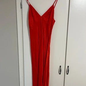Röd  långklänning - Elegant röd långklänning med smala axelband och v-ringning. Perfekt för festliga tillfällen med sin stilrena design och figurnära passform.