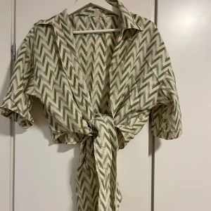 Mönstrad blus med knytning - Säljer en mönstrad blus med gröna och beige sicksack-ränder. Blusen har korta ärmar och en knytning framtill som ger en snygg detalj. Perfekt för en avslappnad stil.