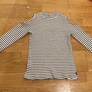 Randig ribbad tröja med öppna axlar - Säljer en snygg randig tröja från KappAhl i storlek 134/140. Tröjan är ribbad med svartvita ränder och har en unik design med öppna axlar. Perfekt för en stilren look!
