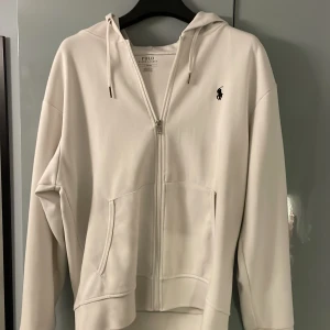 Vit hoodie från Polo Ralph Lauren - Säljer en stilren vit hoodie från Polo Ralph Lauren med dragkedja och klassisk logga på bröstet. Perfekt för en avslappnad look. Den har en bekväm passform och är tillverkad i mjukt material.