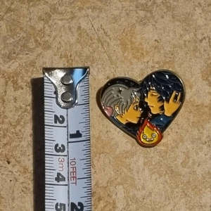 Två emaljpins med motiv från animerad film - Säljer två färgglada emaljpins. Den ena är hjärtformad med två karaktärer och en liten eldfigur. Den andra föreställer en söt eldfigur med vedträ. Perfekta för att dekorera väskor eller jackor!