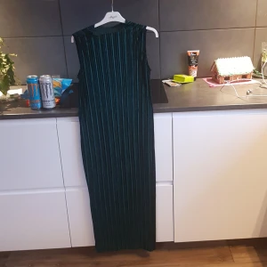 Grön långklänning med ränder - Elegant grön långklänning med vertikala ränder. Klänningen är ärmlös och har en stilren design som passar perfekt för festliga tillfällen. Materialet ser ut att vara mjukt och följsamt. Är plisserad.