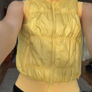 Gul dunväst med dragkedja - Snygg gul dunväst med dragkedja framtill. Perfekt för att hålla värmen under kyliga dagar. Västen har en ribbad kant vid ärmslut och nederkant för extra komfort.