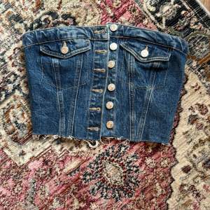 Säljer en cool jeans korsett från Zara med knappar framtill och fransig nederkant. Den har dekorativa fickor och en elastisk del bak för bättre passform. Perfekt för en trendig look!