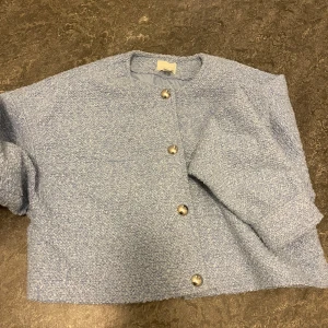 Ljusblå boucléjacka med knappar - Säljer en stilren ljusblå boucléjacka med rund hals och knappar framtill. Jackan har en mjuk textur och är perfekt för att ge en elegant touch till din outfit. Passar bra till både vardag och speciella tillfällen.  ALDRIG ANVÄNT UTAN PRISLAPP!!!