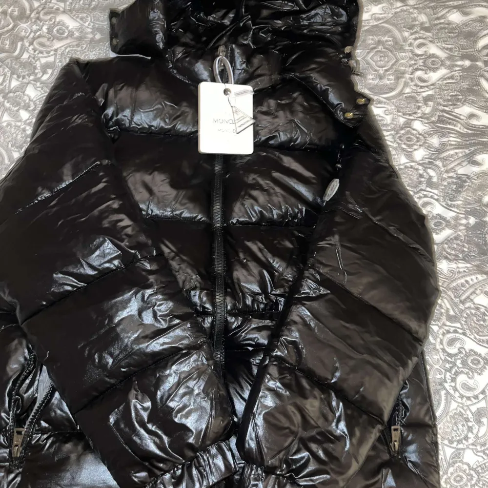 Snygg svart dunjacka från Moncler med glansig finish och dragkedja framtill. Jackan har en klassisk pufferdesign och Moncler-logga på ärmen. Perfekt för kyliga dagar.. Takit.