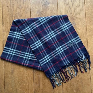 Rutig halsduk från Barbour - Snygg rutig halsduk från Barbour i mörkblått med röda och grå rutor. Perfekt accessoar för att hålla sig varm och stilren under kyliga dagar. Halsduken har fransar i ändarna för en klassisk look.