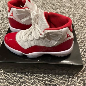 Nike Air Jordan 11 Retro - Snygga Nike Air Jordan 11 Retro i vitt och rött med klassisk design. Skorna har en blank röd yta och vit snörning, perfekt för sneaker-entusiaster. De har en ikonisk Jumpman-logga på sidan och en bekväm passform.