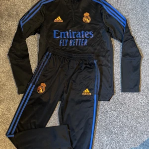 Svart träningsset från Adidas med Real Madrid-logga - Säljer ett svart träningsset från Adidas med Real Madrid-logga. Setet har blå detaljer och dragkedja på tröjan. Perfekt för fotbollsträning eller som sportig vardagsoutfit. Storlek XL men sitter mer som L ( kan passa M också) 