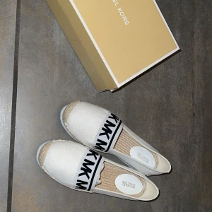 Vita espadrillos från Michael Kors - Snygga vita espadrillos från Michael Kors med svart logotypband. Skorna har en flätad sula och en bekväm, ribbad yttersula. Perfekta för en stilren look. Du får med kartongen.
