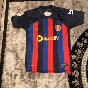 FC Barcelona fotbollströja med Pedri 16 - Snygg FC Barcelona tröja från Nike med Pedri 16 på ryggen. Tröjan har klassiska blå och röda ränder med klubbens emblem på bröstet och Spotify-logga. Kortärmad design perfekt för fotbollsfans. Storlek S