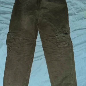 Olivgröna cargopants från Yeezy - Säljer ett par olivgröna cargopants från Yeezy med elastisk midja och dragkedja på fickorna. Byxorna har en avslappnad passform och är perfekta för en casual look. De har även praktiska fickor på sidorna.