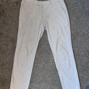 Grå kostymbyxor från Dressmann - Snygga grå kostymbyxor från Dressmann i en klassisk design. Byxorna har en rak passform med midwaist och är perfekta för en stilren look. De har knappar bak och hällor för bälte.