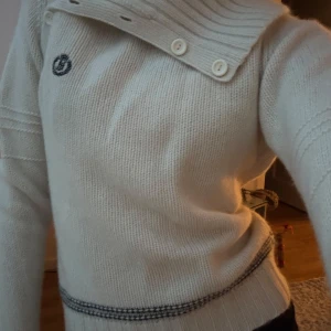 Vit stickad tröja med knappar - Säljer en stilren vit stickad tröja med unika knapplösningar vid kragen. Tröjan har långa ärmar och ribbade detaljer vid ärmslut och nederkant. Perfekt för kyliga dagar.